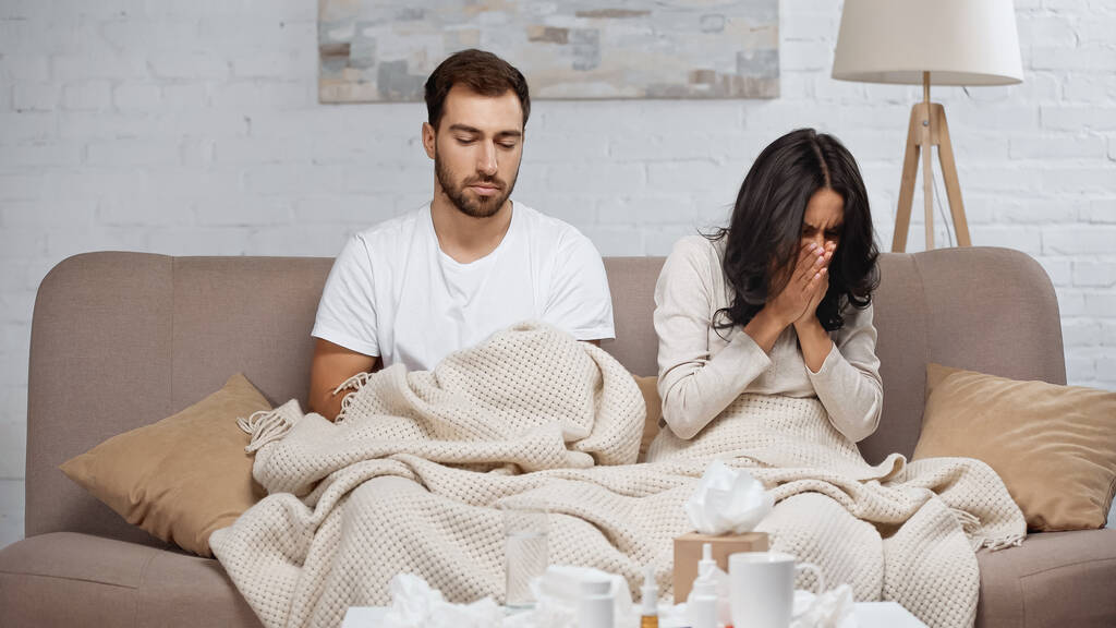 sick woman sneezing near man and bottles with pills on coffee table  - Photo, Image