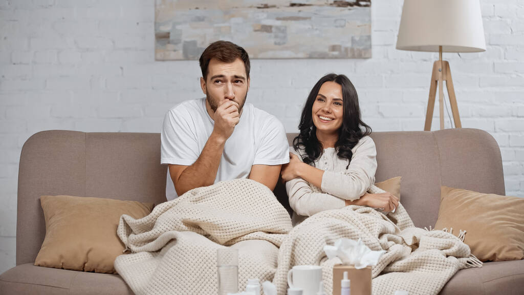 brunette woman smiling near sick boyfriend coughing in living room - Fotoğraf, Görsel