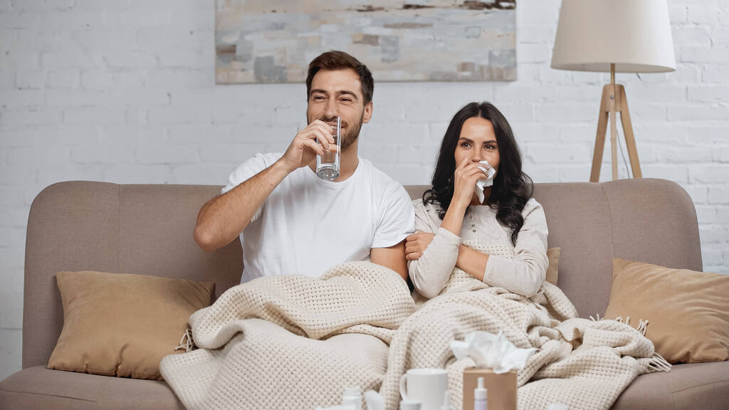 Kranke Frau mit Serviette neben lächelndem Mann trinkt Wasser im Wohnzimmer  - Foto, Bild