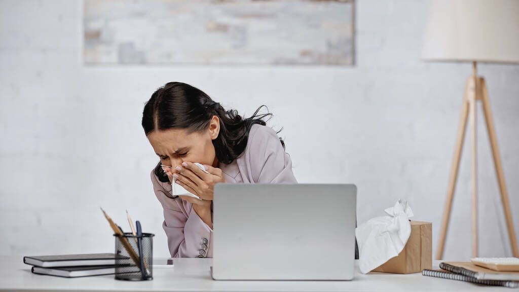 аллергическая деловая женщина чихает в салфетке рядом с ноутбуком на столе - Фото, изображение