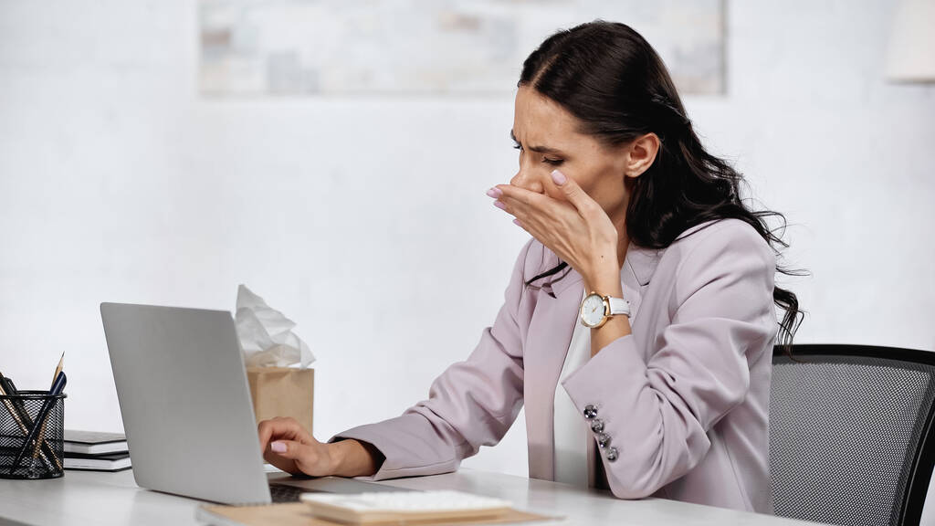 Brünette Geschäftsfrau mit Allergie und rennender Nase neben Laptop auf Schreibtisch - Foto, Bild
