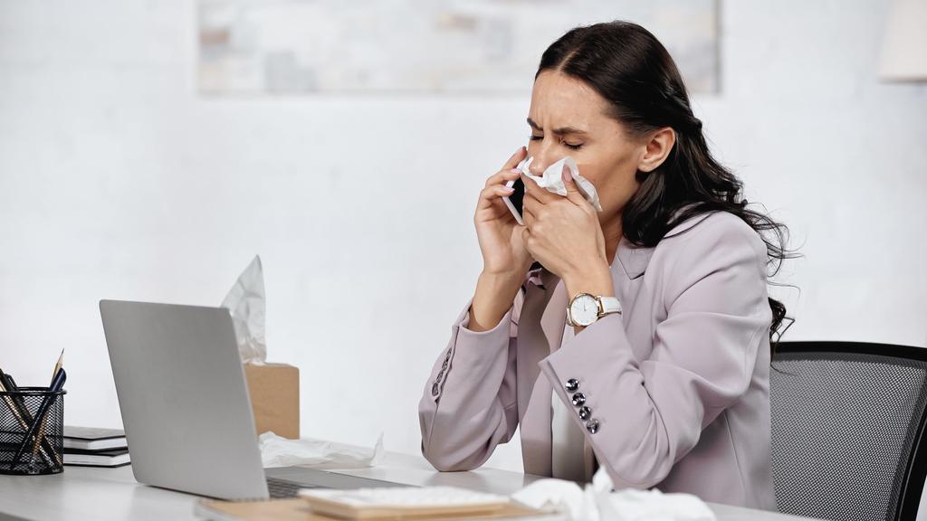 Брюнетка бизнесвумен с аллергией чихает в салфетку во время разговора на смартфоне рядом с ноутбуком на столе - Фото, изображение