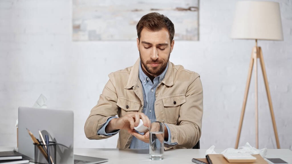 аллергический бизнесмен держит бутылку с таблетками возле ноутбука на столе  - Фото, изображение