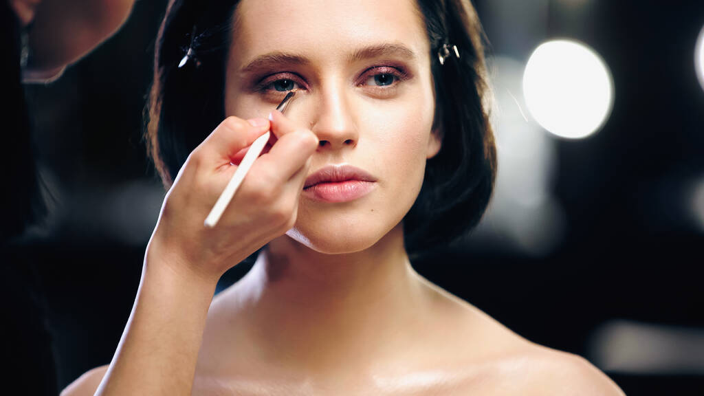 make-up kunstenaar aanbrengen oogschaduw op onderste ooglid van de vrouw met blote schouders - Foto, afbeelding