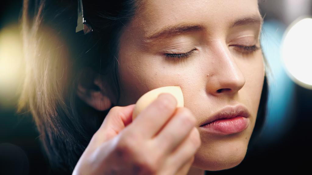 makeup artist applying makeup foundation with cosmetic sponge on face of model with closed eyes - Photo, Image
