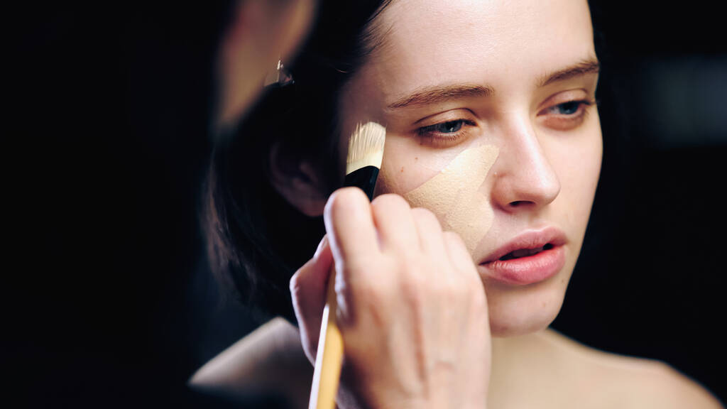 artista de maquiagem segurando escova cosmética ao aplicar a base de maquiagem na pele do jovem modelo  - Foto, Imagem
