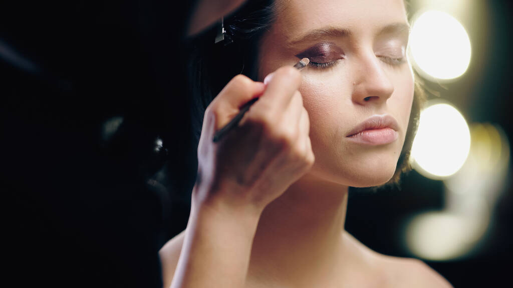 blurred makeup artist applying dark eye shadow with cosmetic brush on eyelids of model with closed eyes - Photo, Image