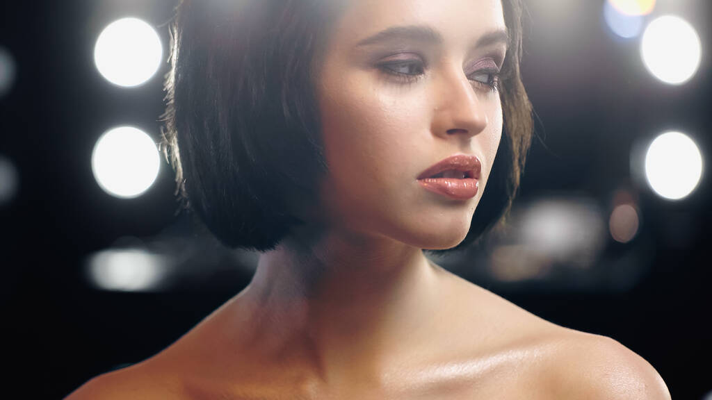 bonita mujer joven con maquillaje y hombros desnudos mirando hacia otro lado con luz de fondo - Foto, Imagen