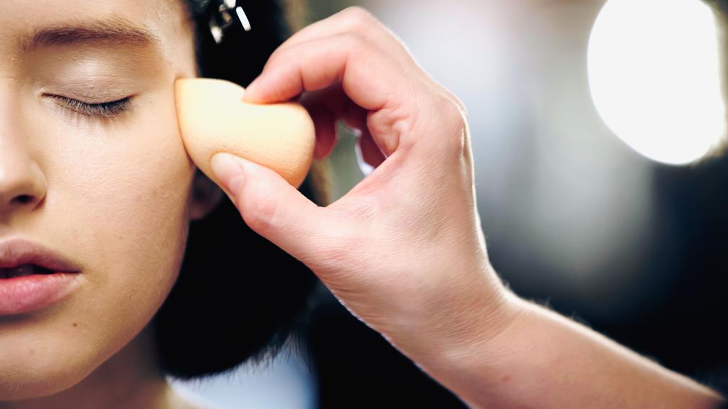 Makyaj sanatçısının kozmetik süngerli kısmi görüntüsü kapalı gözlü bir modele yüz temeli uyguluyor. - Fotoğraf, Görsel