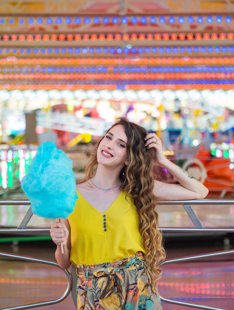 Una hermosa chica caucásica posando con algodón azul de azúcar delante de los coches de choque en un parque - Foto, imagen