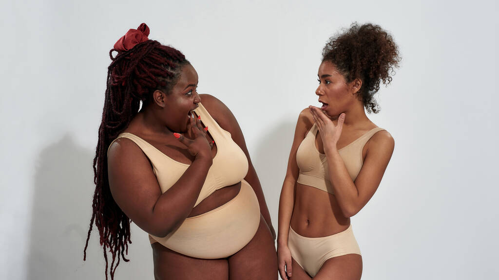 Además de tamaño y delgadas mujeres afroamericanas en ropa interior chismorreando, mirándose emocionalmente, de pie aisladas sobre un fondo gris - Foto, Imagen