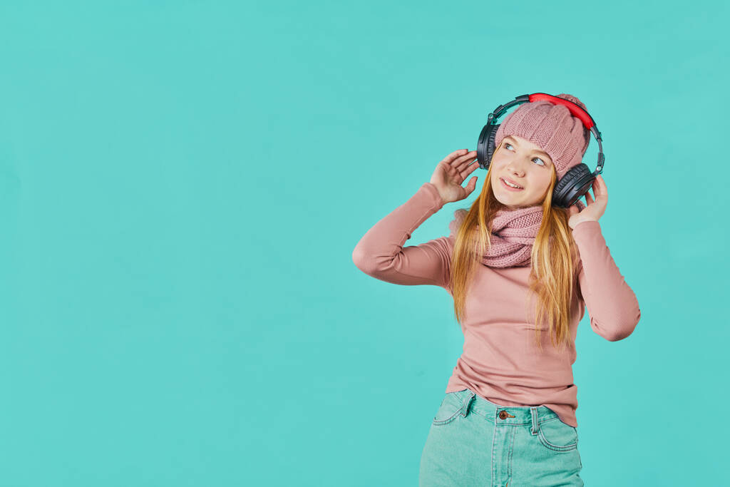 Gyönyörű fiatal nő zenét hallgat fejhallgatóval mosolyog, oldalra néz. Portré érzelmi lány kalap kék háttér. - Fotó, kép