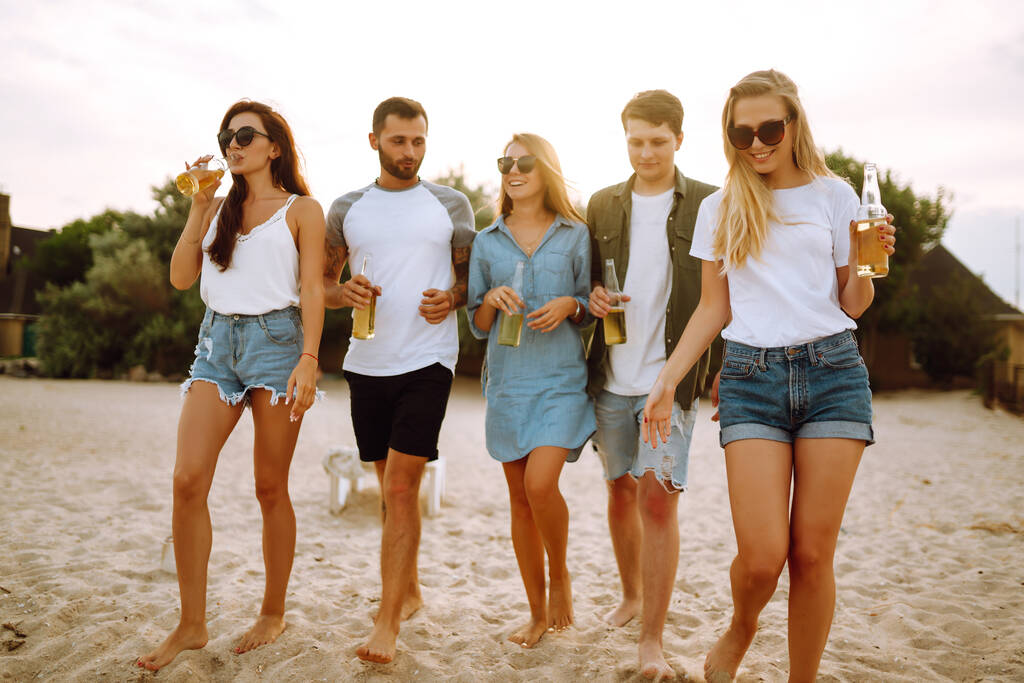Grupo de amigos aplausos y beber cervezas en la playa. Jóvenes amigos que se relajan en el beath haciendo picnic, brindando con cerveza. Concepto de personas, estilo de vida, viajes, naturaleza y vacaciones. - Foto, Imagen
