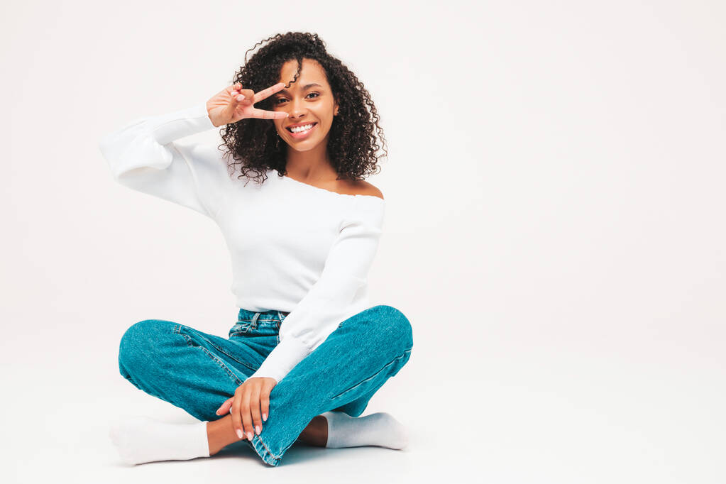 Bella donna nera con riccioli afro acconciatura.Modello sorridente in maglione e jeans alla moda. Sexy donna spensierata seduta su sfondo bianco in studio. Abbronzato e allegro. Mostra segno di pace - Foto, immagini