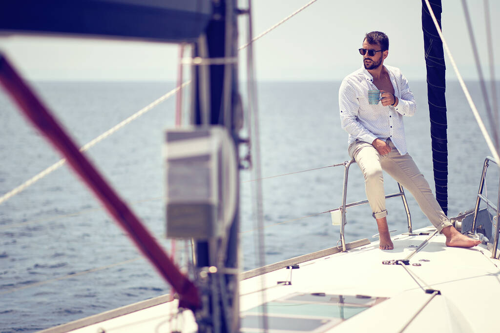 Un joven guapo modelo masculino está disfrutando del sol y el café en una sesión de fotos en un yate en un hermoso día soleado en la orilla del mar. Verano, mar, vacaciones - Foto, imagen