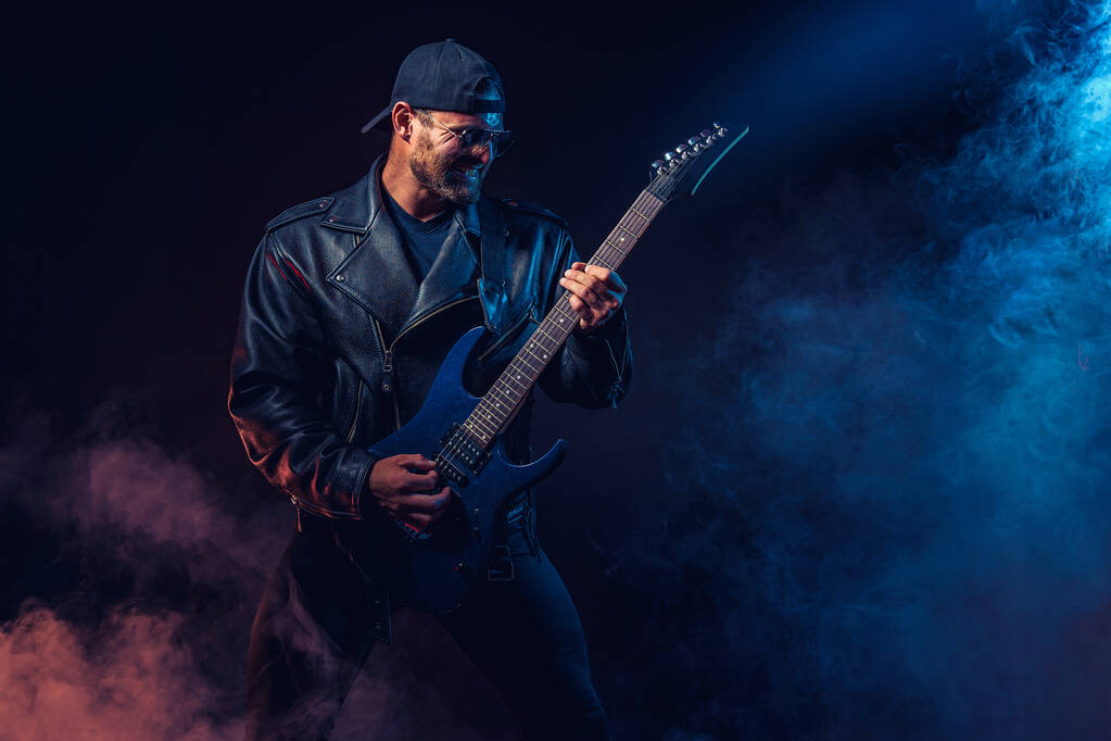 Brutaler bärtiger Heavy-Metal-Musiker in Lederjacke und Sonnenbrille spielt E-Gitarre. Aufnahme in einem Studio auf dunklem Hintergrund mit Rauch - Foto, Bild