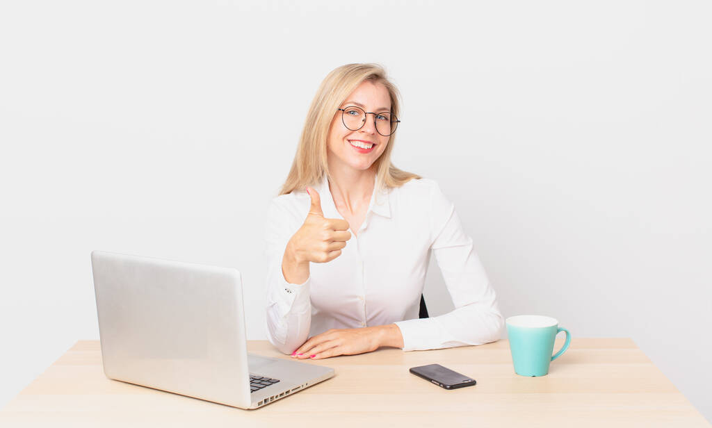 blonde jolie femme jeune femme blonde se sentant fière, souriant positivement avec les pouces levés et travaillant avec un ordinateur portable - Photo, image