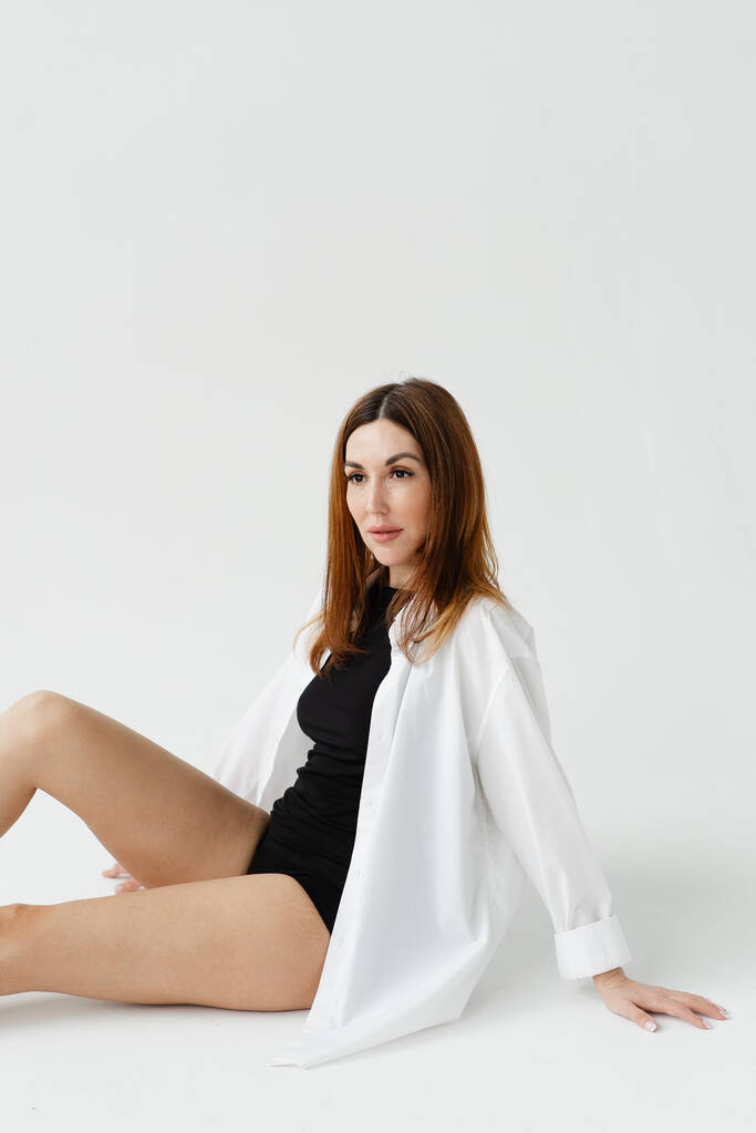 hermosa chica con las piernas desnudas en una camisa blanca se sienta en el suelo sobre un fondo liso - Foto, imagen