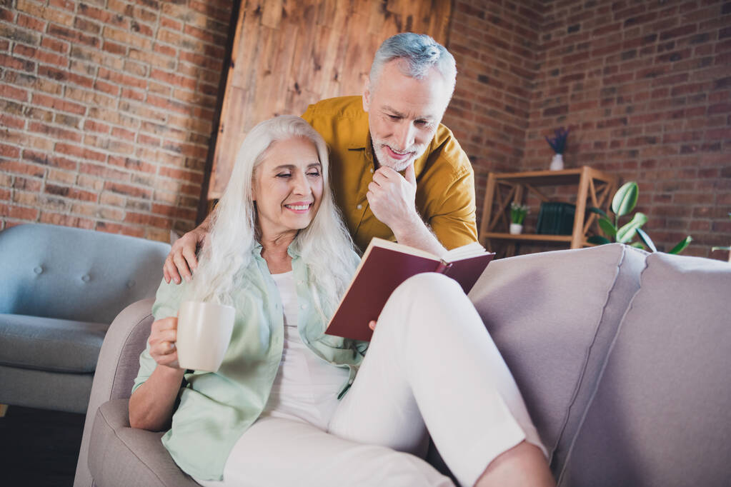 Фото привабливого позитивного щасливого старого чоловіка і дружини читають книгу напій кавовий кухоль в приміщенні будинку
 - Фото, зображення