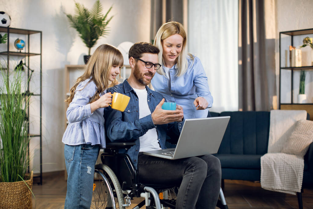 Женщина и ребенок стоят рядом с мужчиной в инвалидной коляске с ноутбуком - Фото, изображение