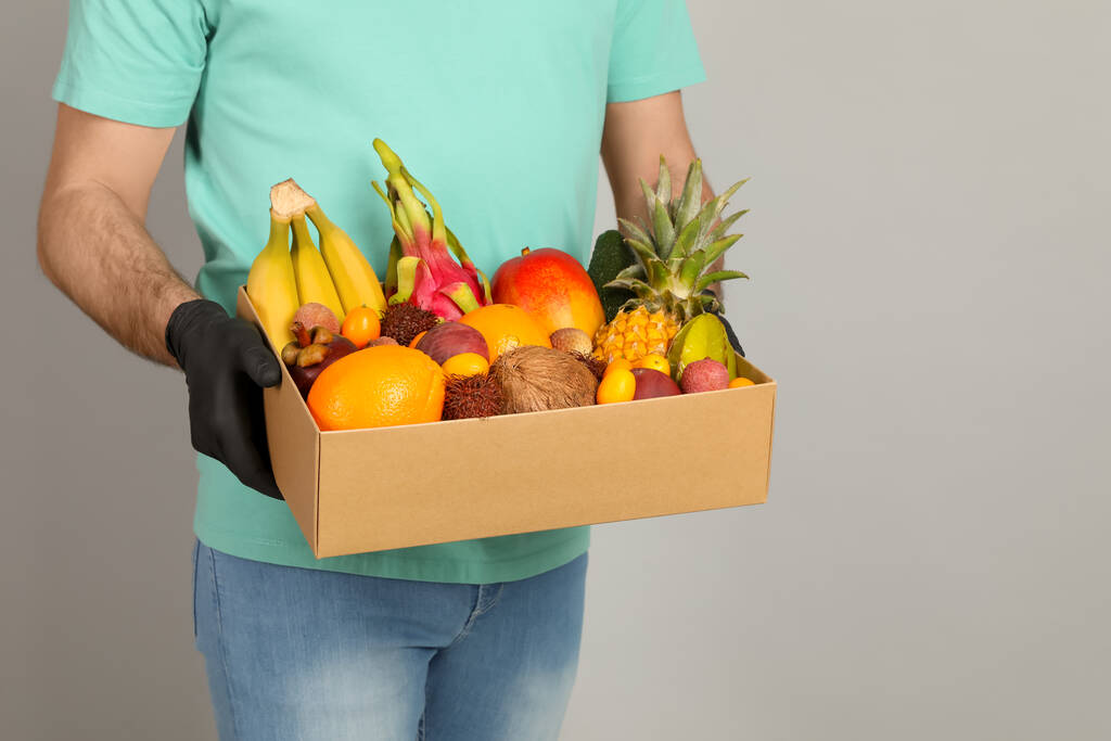 Кур'єр тримає коробку з різноманітними екзотичними фруктами на сірому фоні, крупним планом
 - Фото, зображення