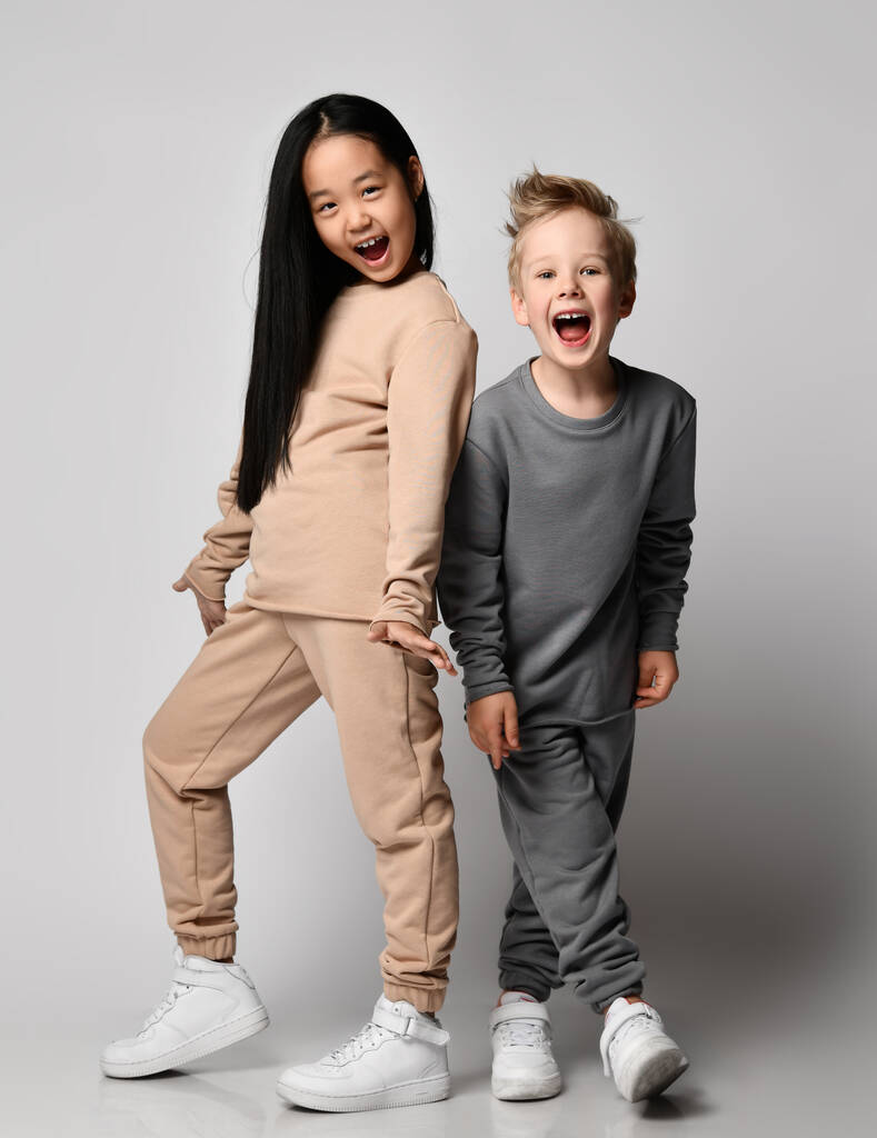 Дети в уютной хлопчатобумажной спортивной одежде играют вместе. Блондин мальчик и брюнетка девочка в брюках и толстовках стоит рядом - Фото, изображение