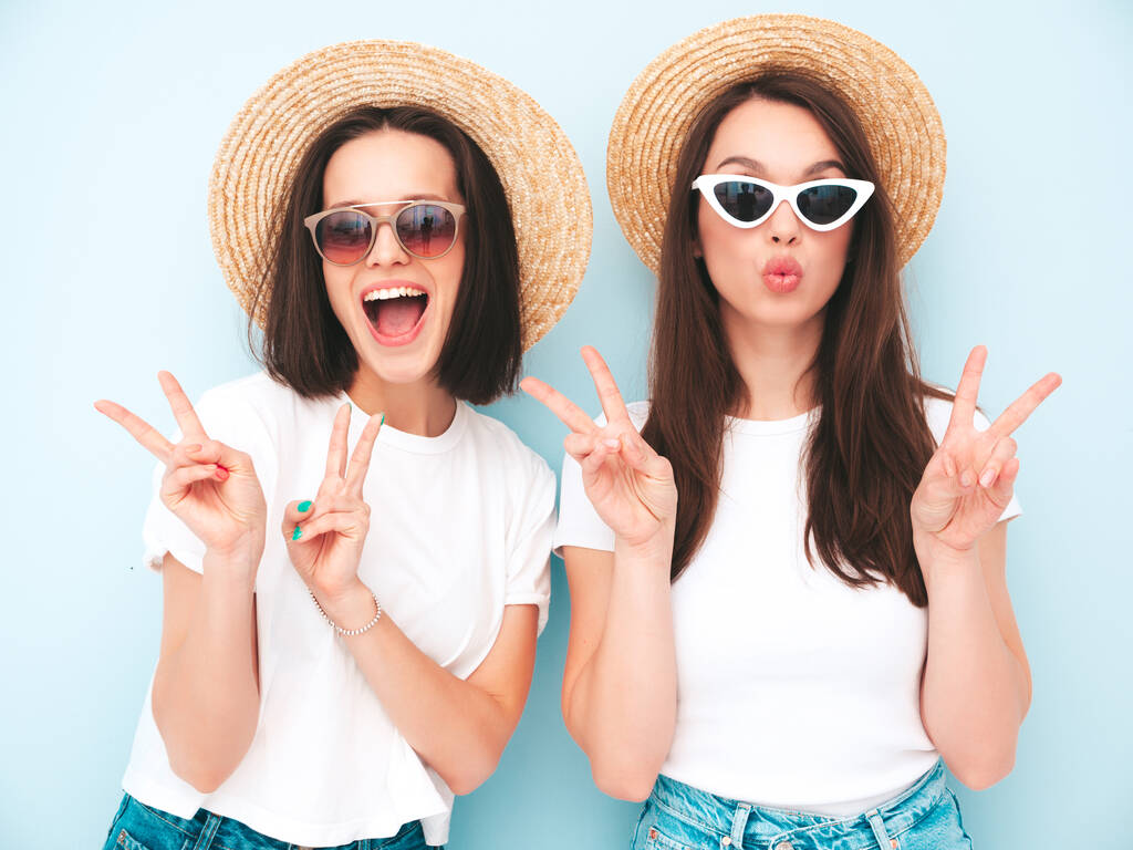 Zwei junge schöne lächelnde Hipsterinnen in trendigen sommerlichen weißen T-Shirts und Jeanskleidern. Sexy unbeschwerte Frauen posieren in der Nähe hellblauer Wände im Studio.Positive Models mit Hüten. Zeigen Friedenszeichen - Foto, Bild