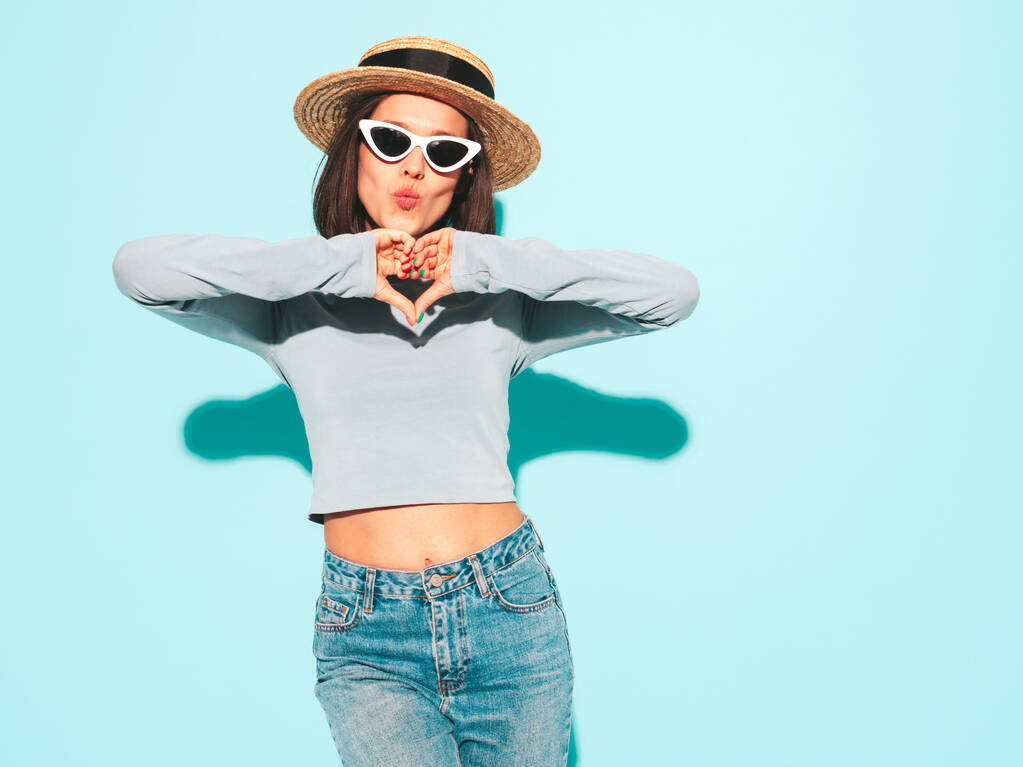Portré fiatal gyönyörű mosolygós nő divatos nyári hipster ruhákat. Szexi gondtalan nő pózol a kék fal mellett a stúdióban. Pozitív modell, aki napszemüvegben és kalapban szórakozik. Szívet jelez - Fotó, kép