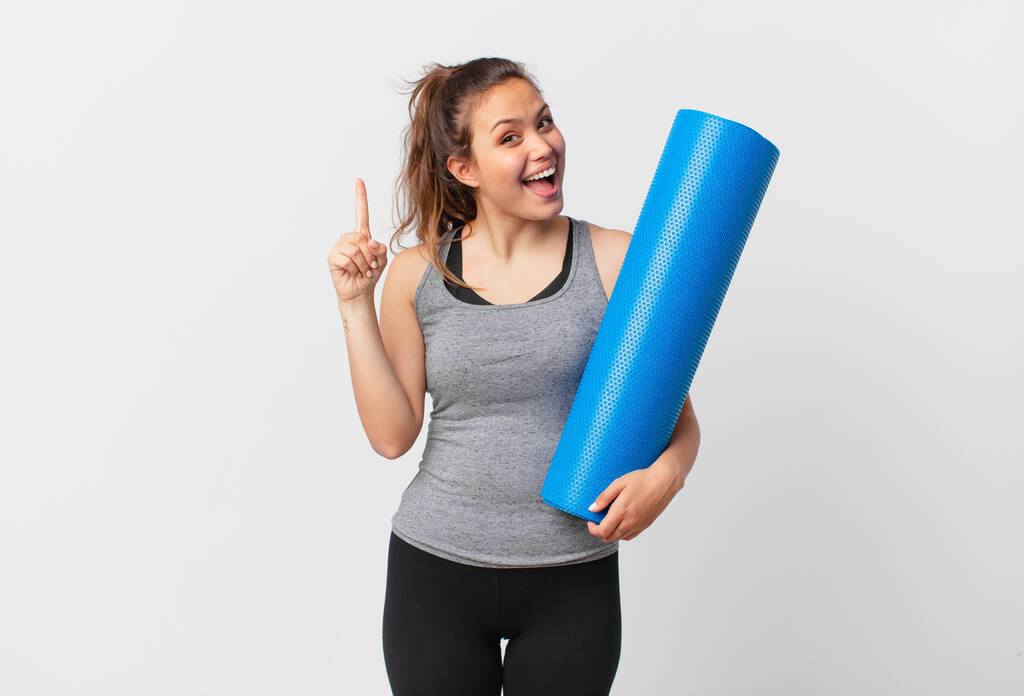 young pretty woman feeling like a happy and excited genius after realizing an idea and holding a yoga mat - Photo, Image