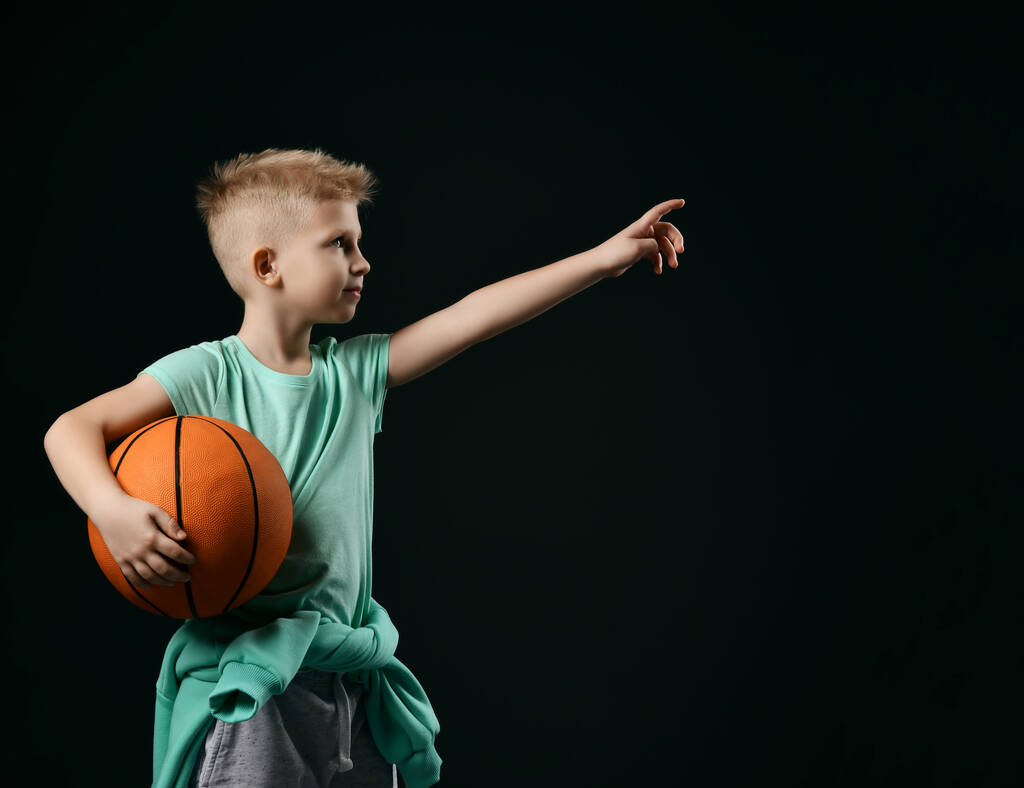 Lächelndes blondes Knabenkind in sportgrünem T-Shirt und Hose stehend, Basketball in den Händen, mit dem Finger nach oben zeigend - Foto, Bild