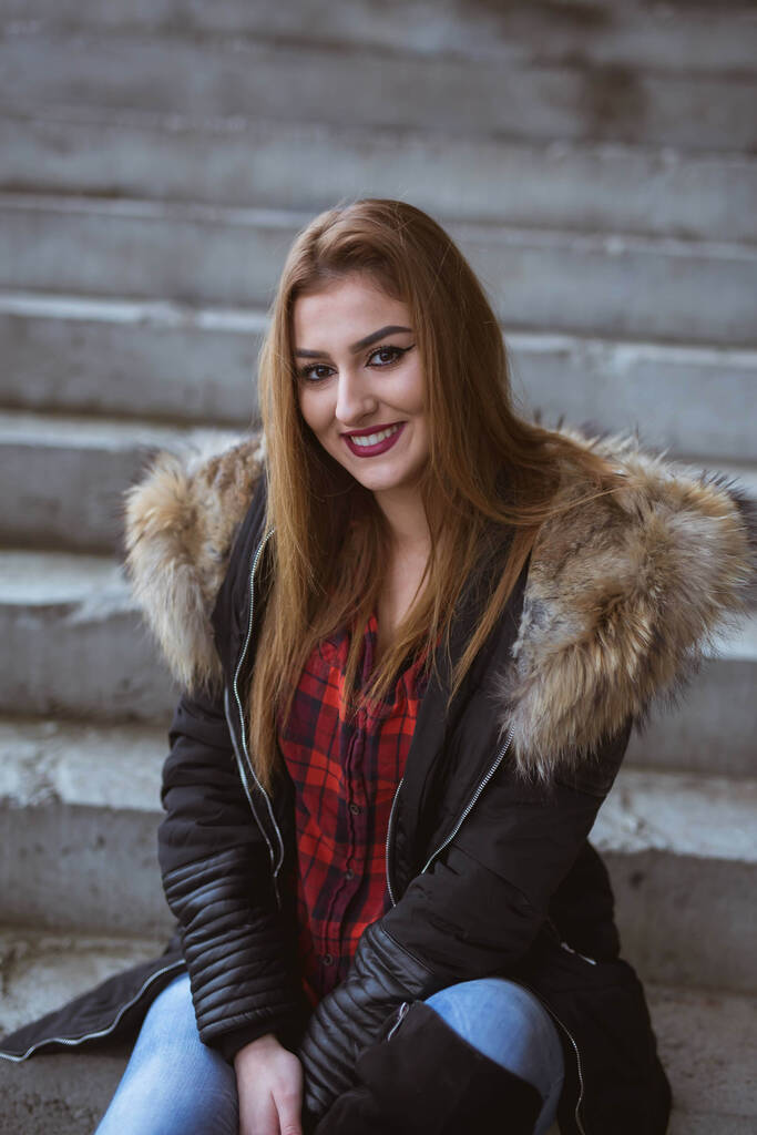 Молода жінка з білявим волоссям в пальто сидить на сходах і посміхається
 - Фото, зображення