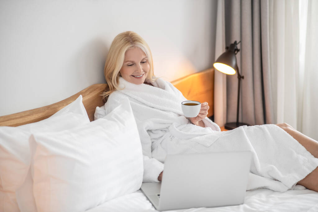 Una mujer de blanco acostada en la cama y mirando algo en el portátil - Foto, Imagen