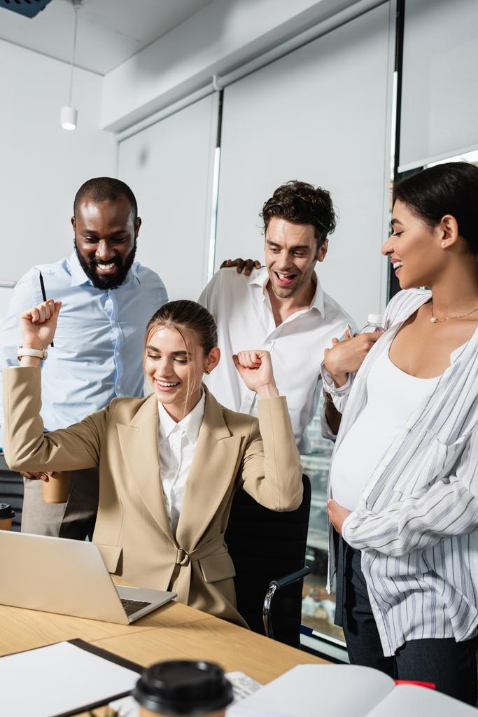 excited businesswoman showing win gesture near laptop and multiethnic colleagues - Photo, Image