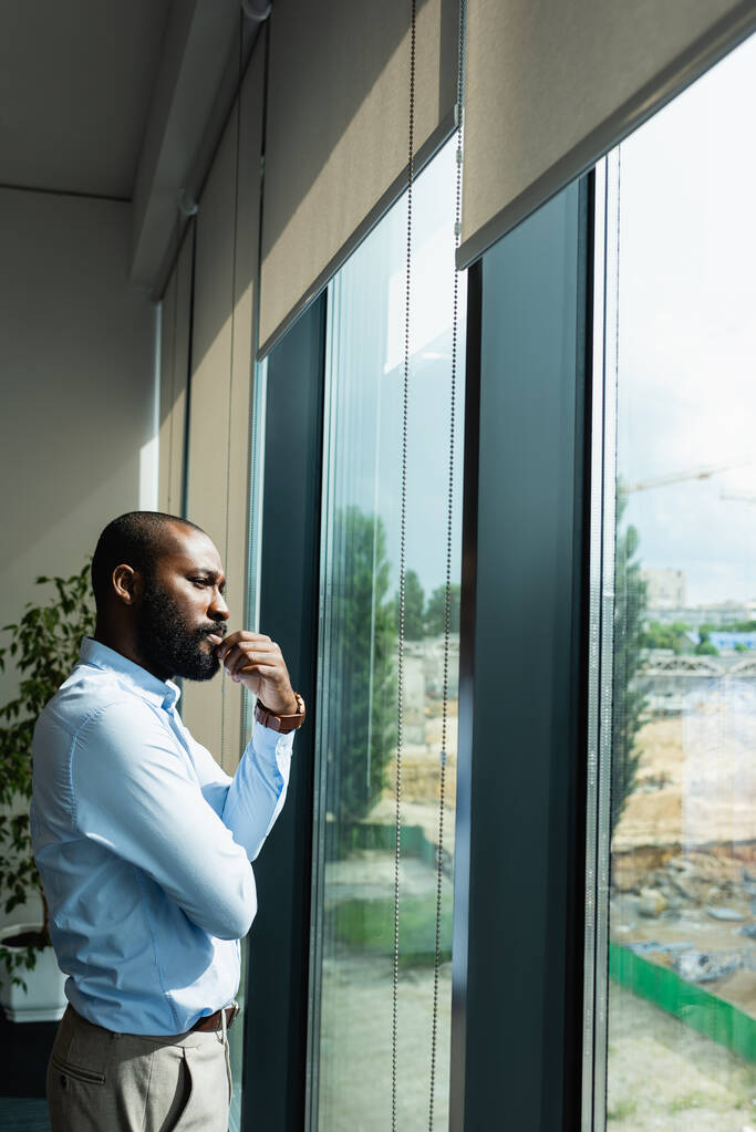 figyelmes afro-amerikai üzletember félrenéz, miközben az ablak mellett áll. - Fotó, kép