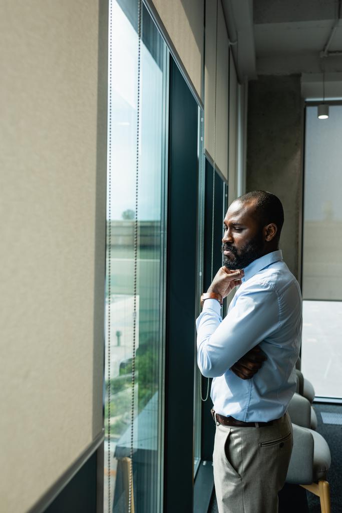 σοβαρός Αφροαμερικάνος επιχειρηματίας που σκέφτεται κοντά σε παράθυρο στο γραφείο - Φωτογραφία, εικόνα