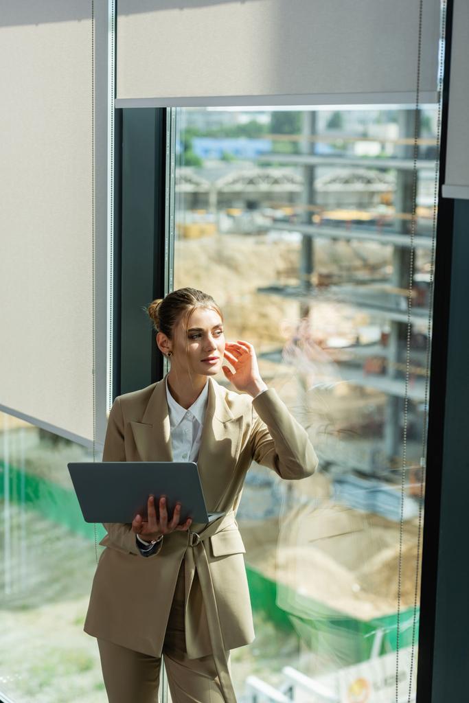 dreamy businesswoman holding laptop while looking away through window in office - Photo, Image