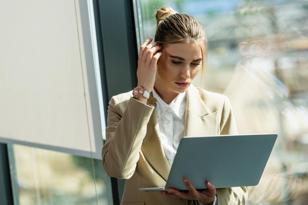 pensativa mujer de negocios fijar el pelo mientras mira el ordenador portátil en la oficina - Foto, Imagen