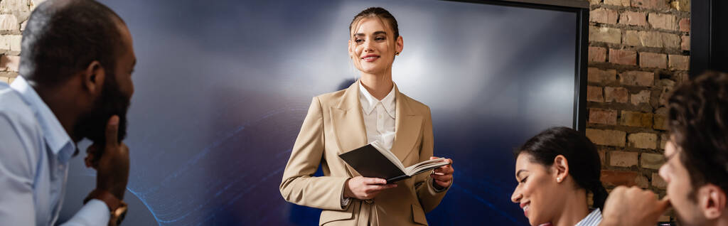 donna d'affari sorridente in piedi con notebook vicino a colleghi interrazziali, banner - Foto, immagini