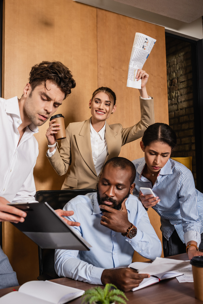 excited businesswoman holding newspaper near thoughtful interracial colleagues - Photo, Image
