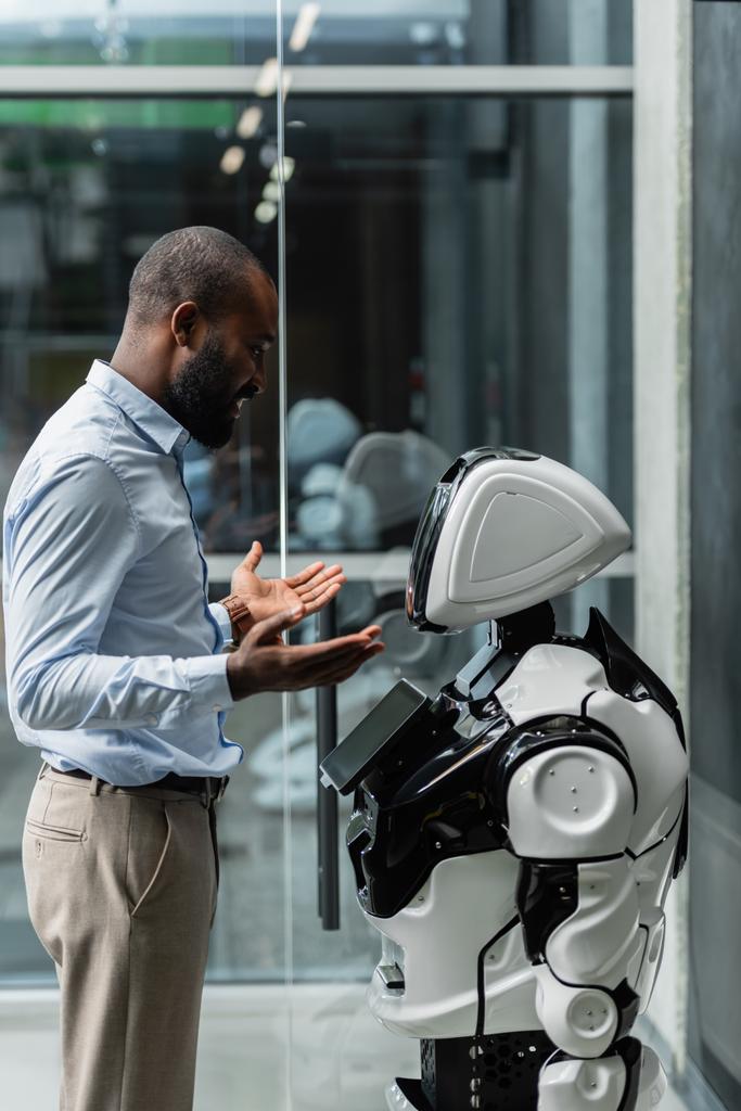 glimlachen Afrikaans amerikaanse zakenman gebaren in de buurt van robot in het kantoor - Foto, afbeelding