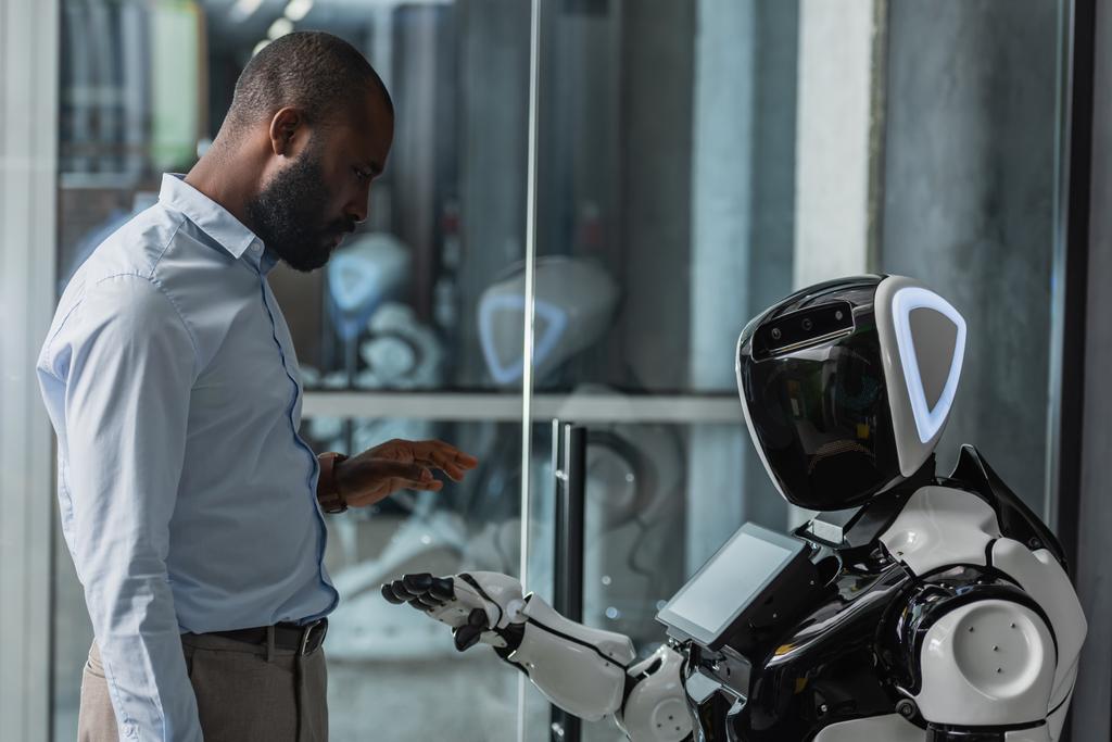 Afrykański amerykański biznesmen stojący obok humanoidalnego robota w biurze - Zdjęcie, obraz