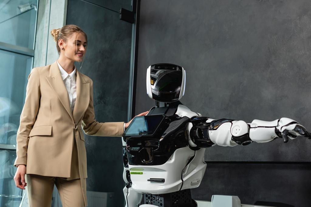 χαρούμενη επιχειρηματίας που αγγίζει ανθρωποειδές ρομπότ στο γραφείο - Φωτογραφία, εικόνα