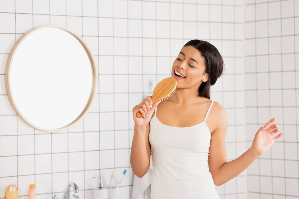 giovane donna afroamericana in abiti bianchi che canta con spazzola per capelli e gesticola con mano in bagno  - Foto, immagini