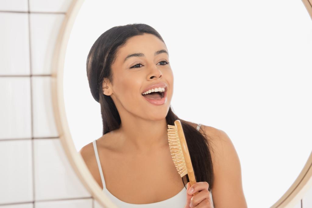junge afrikanisch-amerikanische Frau singt mit Haarbürste in der Nähe von Spiegel im Badezimmer - Foto, Bild