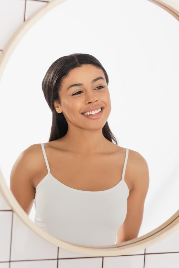 Beyaz kolsuz tişörtlü, banyoda aynaya bakan gülümseyen Afro-Amerikan bir kadın. - Fotoğraf, Görsel
