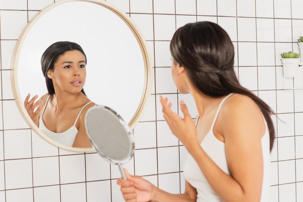 若いアフリカ系アメリカ人の女性が鏡を見てバスルームで顔の近くに手を取り - 写真・画像