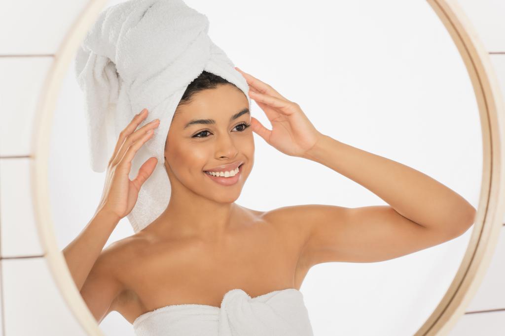 joven mujer afroamericana envuelta en toallas cogidas de la mano cerca de la cabeza y mirando en el espejo en el baño - Foto, imagen