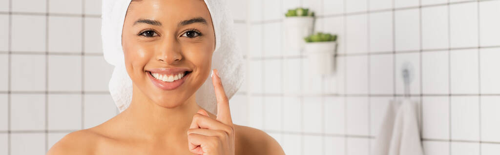 giovane donna afroamericana che applica crema sul viso con dito in bagno, banner - Foto, immagini