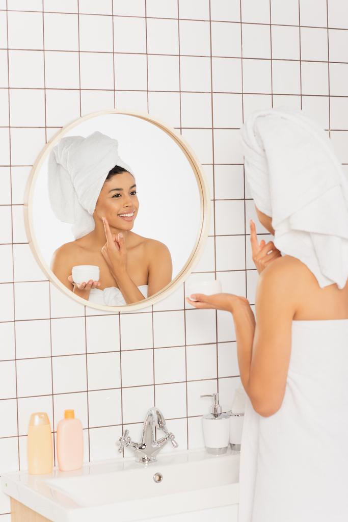 νεαρή Αφροαμερικανή γυναίκα τυλιγμένη σε πετσέτες απλώνοντας κρέμα στο πρόσωπο κοντά στον καθρέφτη στο μπάνιο - Φωτογραφία, εικόνα