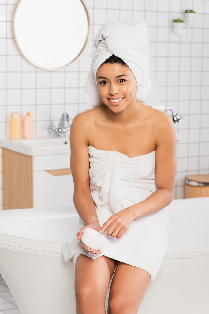 usmívající se mladá africká americká žena sedí na vaně a drží sklenici se smetanou v koupelně - Fotografie, Obrázek